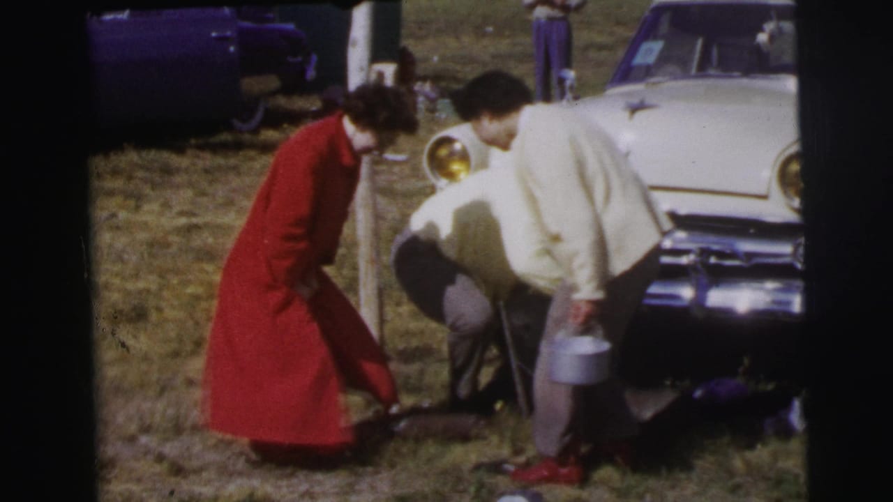 A woman in red and a man in a light sweater bend toward gear on the grass beside a white car in Le Mans, France.