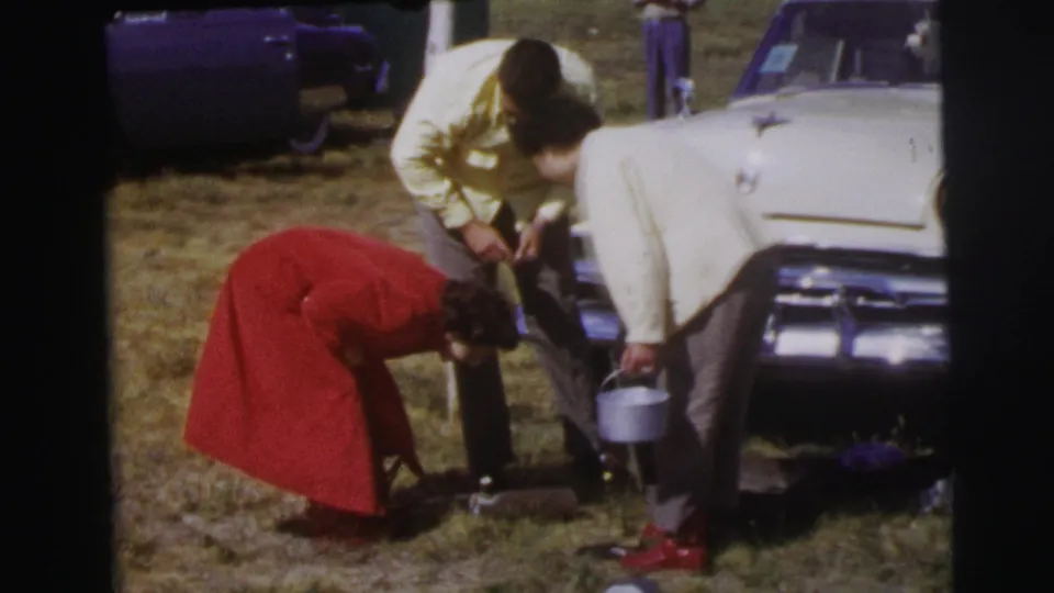 Three adults cluster beside a white car, with one figure in a red coat reaching toward items on the grass.