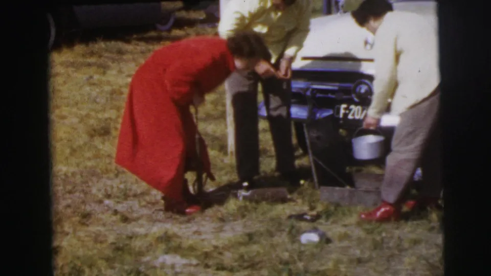 The woman in red bends over the grass as men stand nearby with a bucket and tools beside the front of a white car.