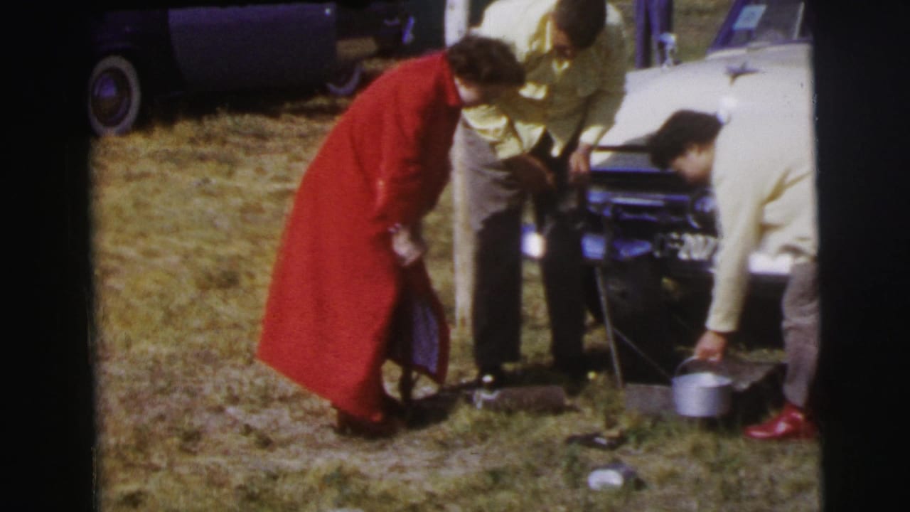 In Le Mans home-movie footage from 1954, a woman in a red coat tends items on the grass beside a pale classic car as two men look down.