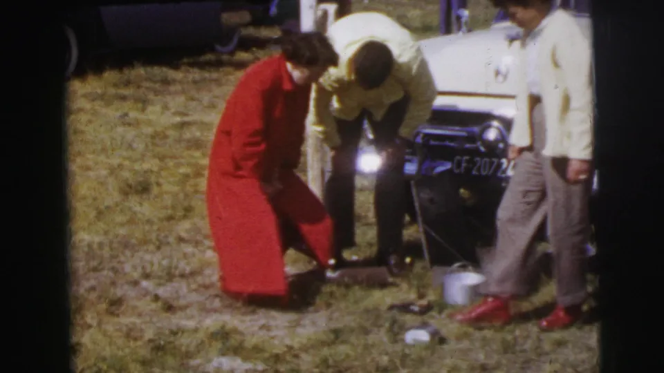 The woman in red crouches by a pot on the grass as men in light shirts stand beside a pale classic car in Le Mans, France.