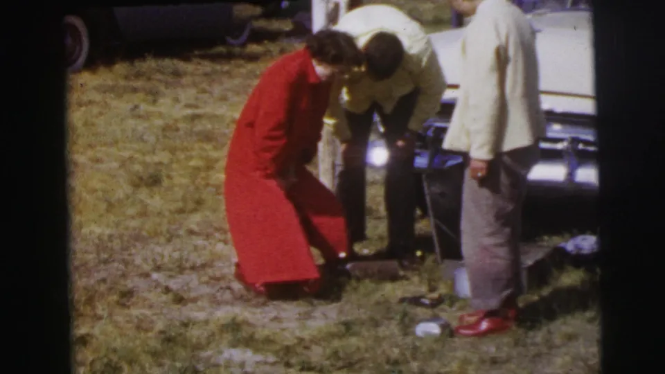 A woman in a bright red coat stands near cooking items while two men face the front of a pale classic car in the Le Mans field scene.