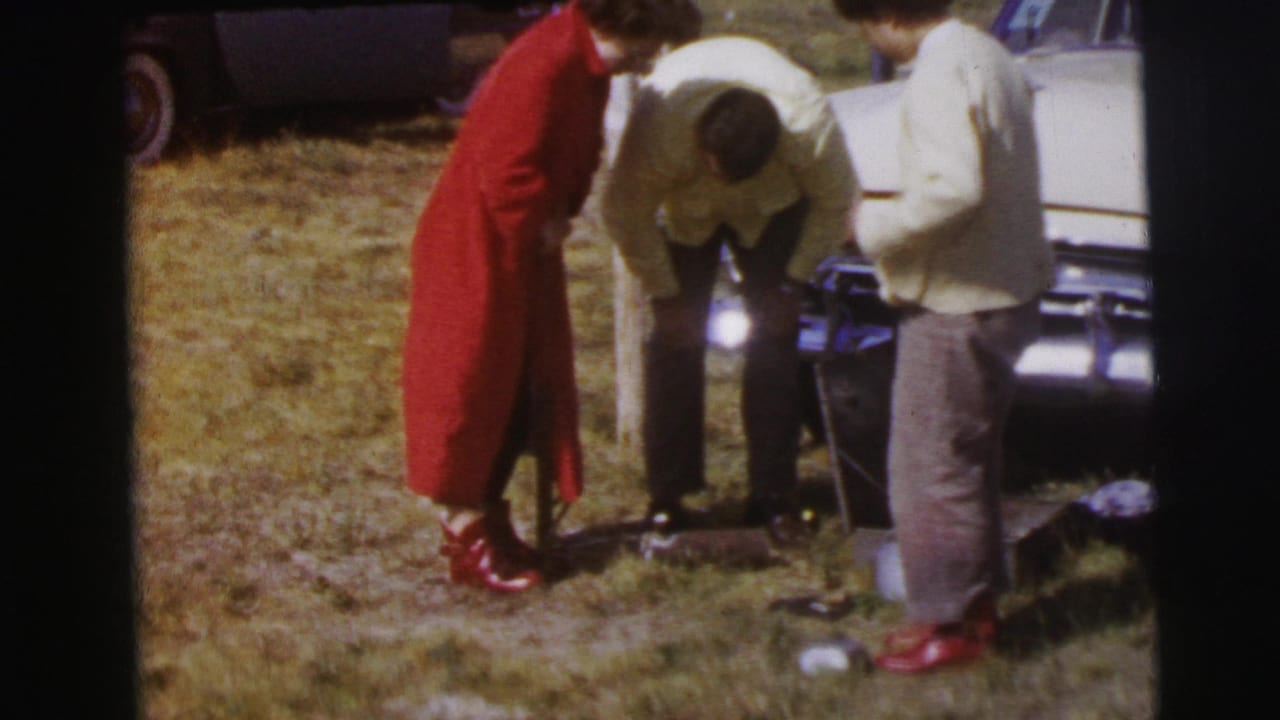 At Le Mans, France in 1954, three people stand beside a white car on grass, including a person in red leaning down near equipment or the ground.