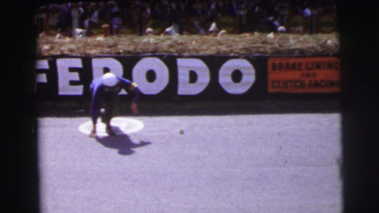 At Le Mans, France in 1954, a driver in a blue suit and white helmet crouches on the asphalt near a Ferodo sign and spectators behind hay bales.