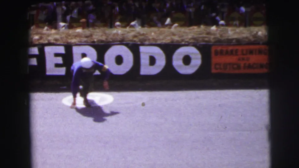 At Le Mans, France in 1954, a driver in a blue suit and white helmet crouches on the asphalt near a Ferodo sign and spectators behind hay bales.