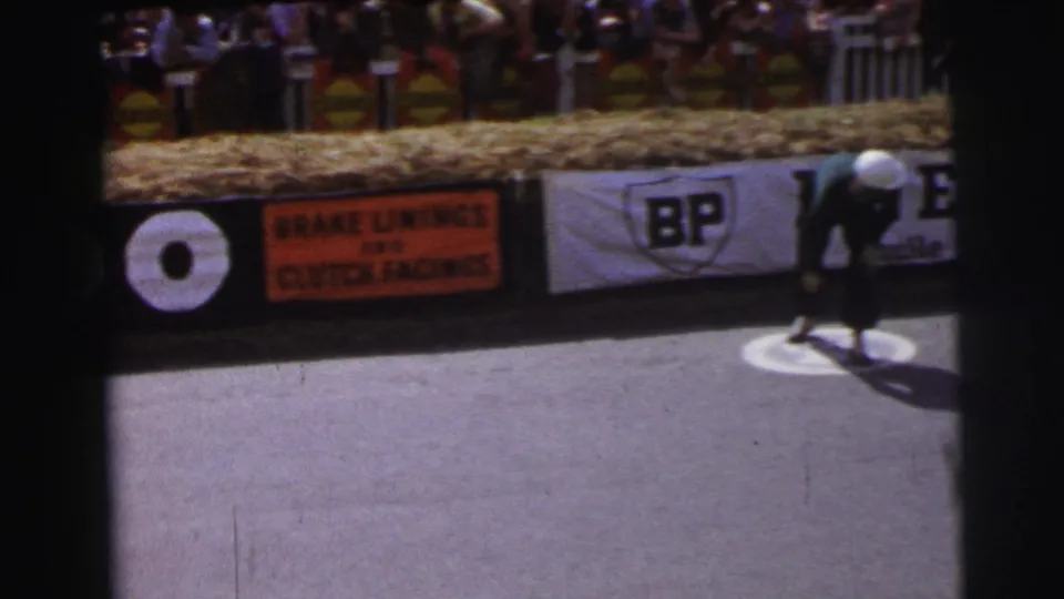 At Le Mans, France in 1954, a helmeted driver bends low on the track in front of Ferodo lettering, a BP banner, and an orange brake-lining advertisement.