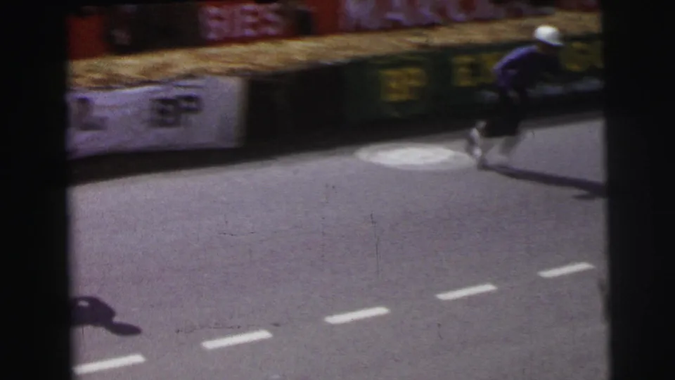 At Le Mans, France in 1954, a driver in a blue racing suit runs across the asphalt past hay bales and partially blurred trackside advertising.