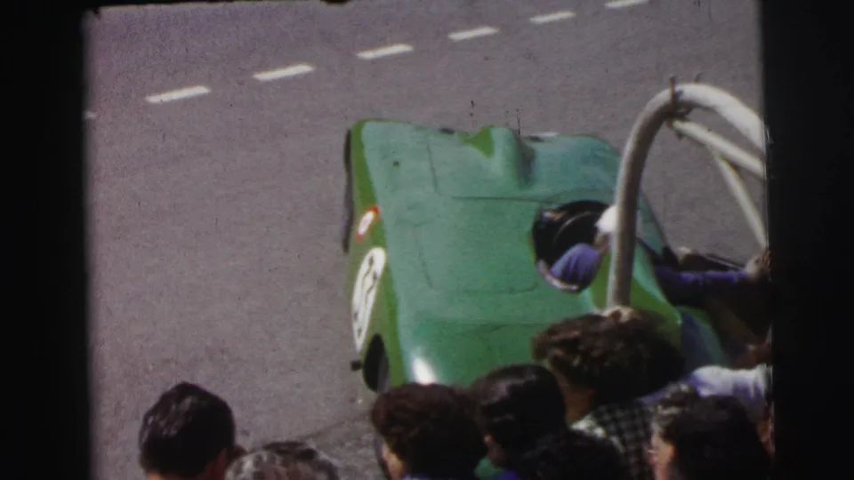 1954 Le Mans home-movie frame showing a person in purple entering a green open race car as the crowd watches.