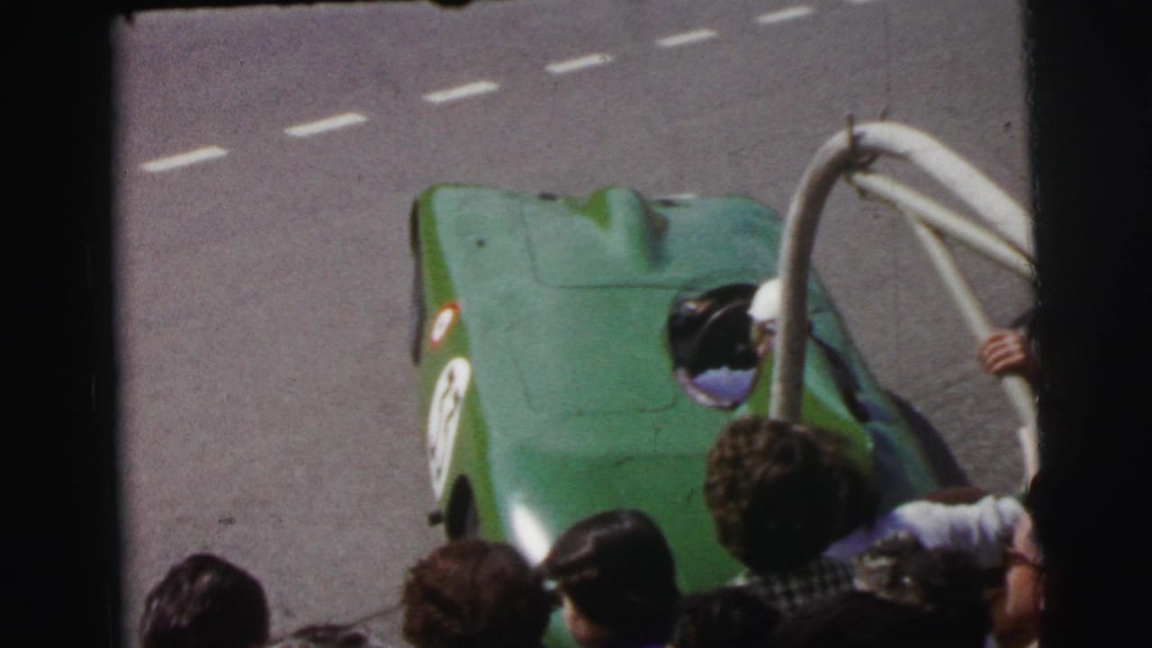 At Le Mans in 1954, a green open-cockpit race car passes below a crowd and roadside railing on the track.