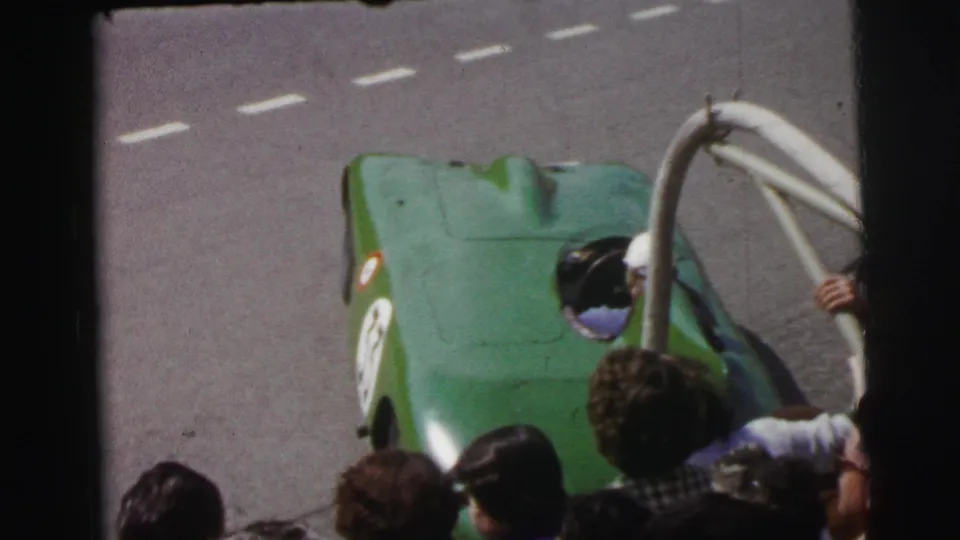At Le Mans in 1954, a green open-cockpit race car passes below a crowd and roadside railing on the track.