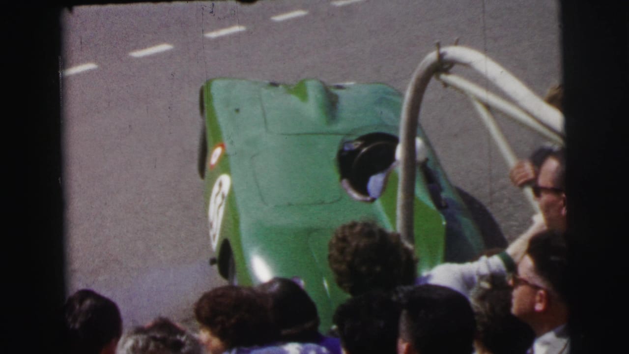 A green open-cockpit race car at the 1954 Le Mans race passes spectators gathered beside the track.
