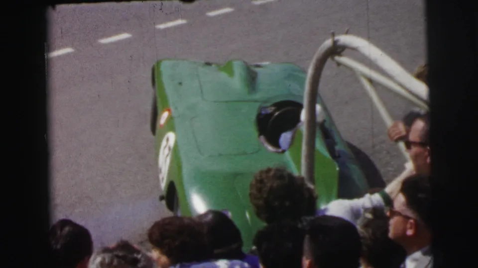 A green open-cockpit race car at the 1954 Le Mans race passes spectators gathered beside the track.