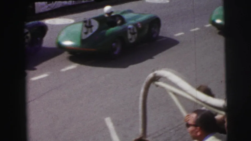 A blue-green open-cockpit race car passes by spectators at the 1954 Le Mans race, with roadside railing in front.