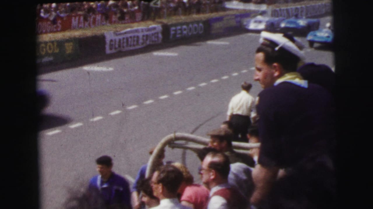 At the 1954 Le Mans setting, spectators in sailor-style caps look toward a blue racing car on the circuit.