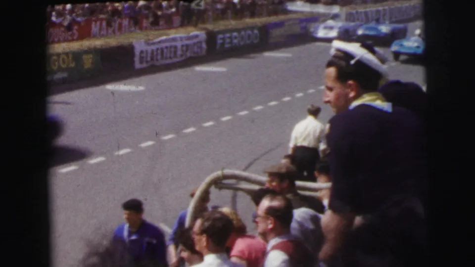 At the 1954 Le Mans setting, spectators in sailor-style caps look toward a blue racing car on the circuit.