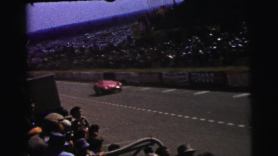 A red sports car travels along the Le Mans track while spectators watch from a shaded foreground position.