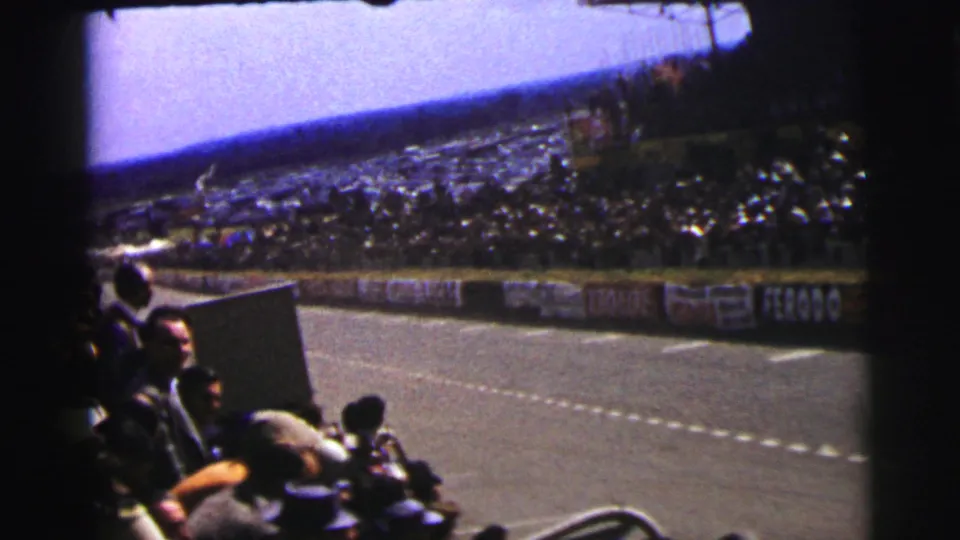 The Le Mans circuit is seen from a spectator viewpoint, with crowds, barriers, and trackside signs framing the empty stretch of road.