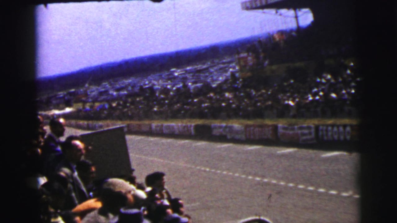 Spectators watch a race track from beside a crowded grandstand under bright sky.
