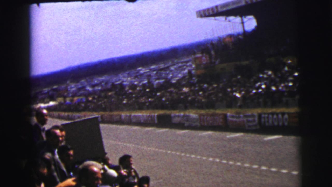 Le Mans spectator view with seated crowds, foreground viewers, track barriers, and period advertising along the circuit.