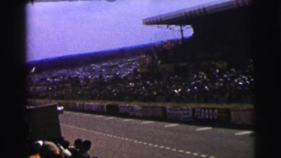 Soft 8mm frame of the Le Mans circuit area, with packed stands, trackside barriers, and spectators seen from a tilted crowd viewpoint.