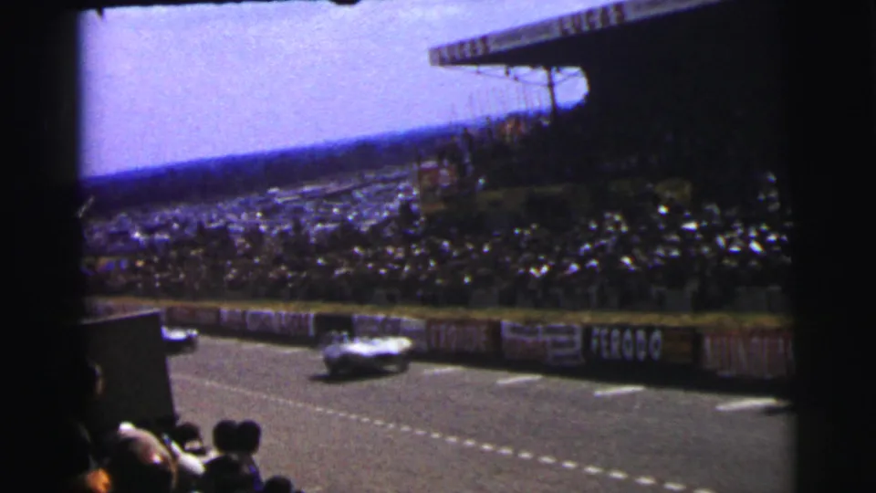 1954 Le Mans frame with a low race car passing the spectator stands, advertising boards, and foreground crowd.