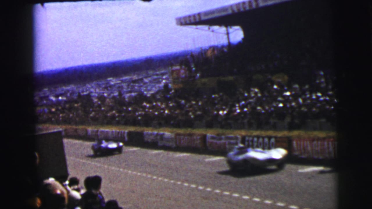 1954 Le Mans 8mm frame showing racing cars speeding past a crowd, track barriers, and period advertising banners.