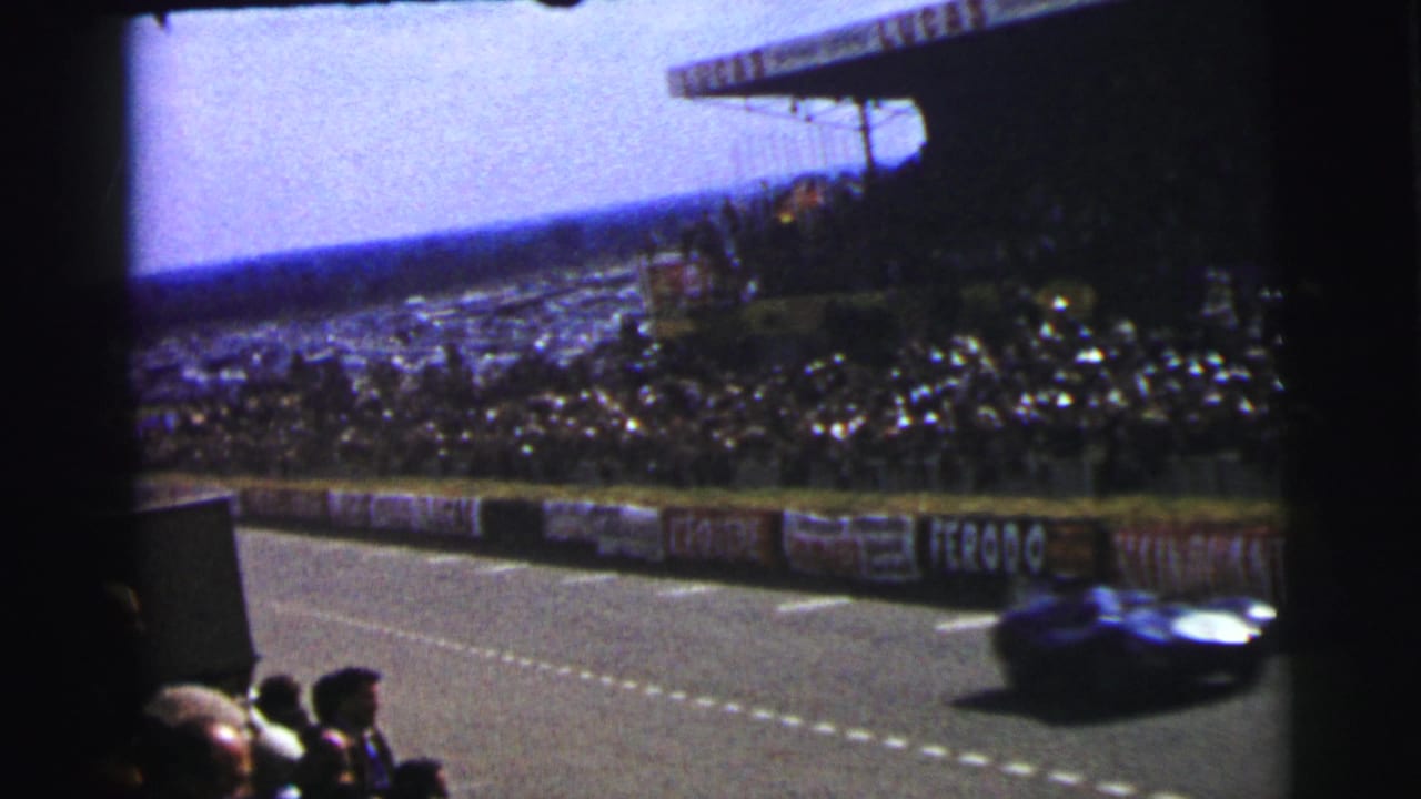 1954 Le Mans 8mm frame with a blue racing car moving past foreground spectators, barriers, and packed stands.