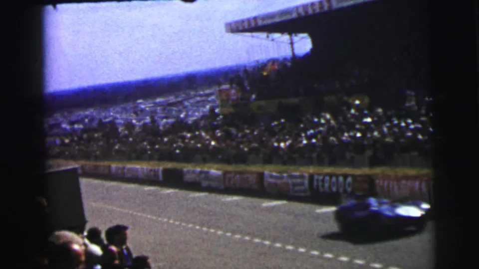 1954 Le Mans 8mm frame with a blue racing car moving past foreground spectators, barriers, and packed stands.