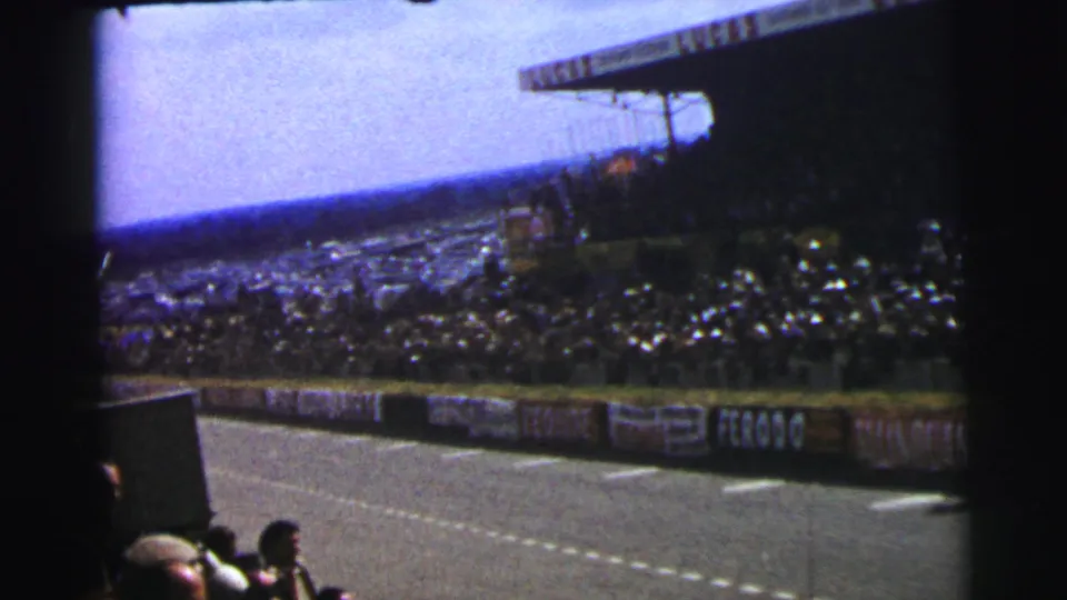 1954 Le Mans 8mm frame showing the track, foreground spectators, distant crowd, and period advertising along the barrier.