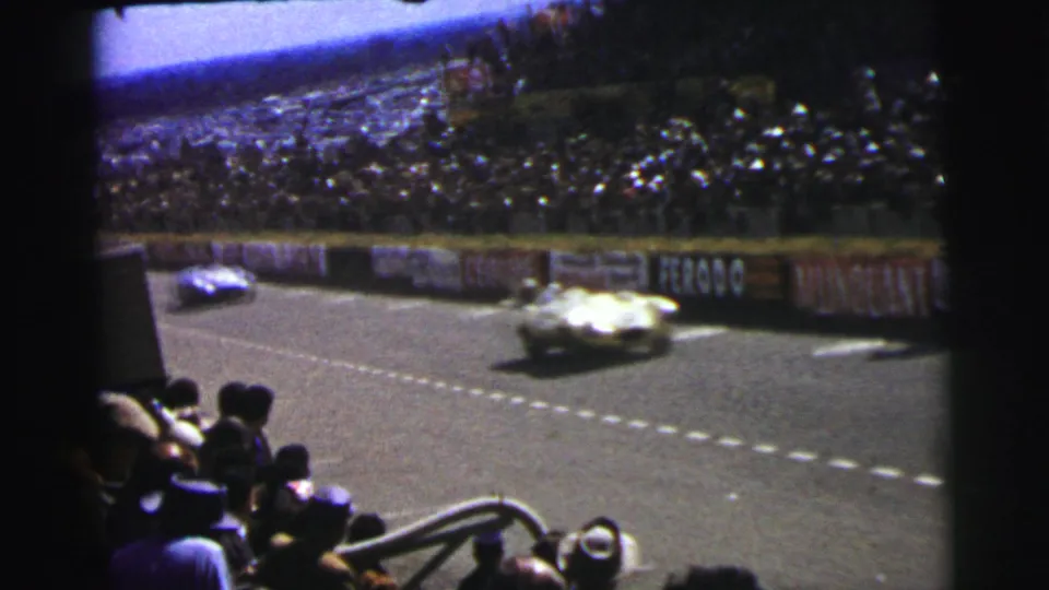 1954 Le Mans 8mm frame with two racing cars on the course, spectators below, and brand signage along the barrier.