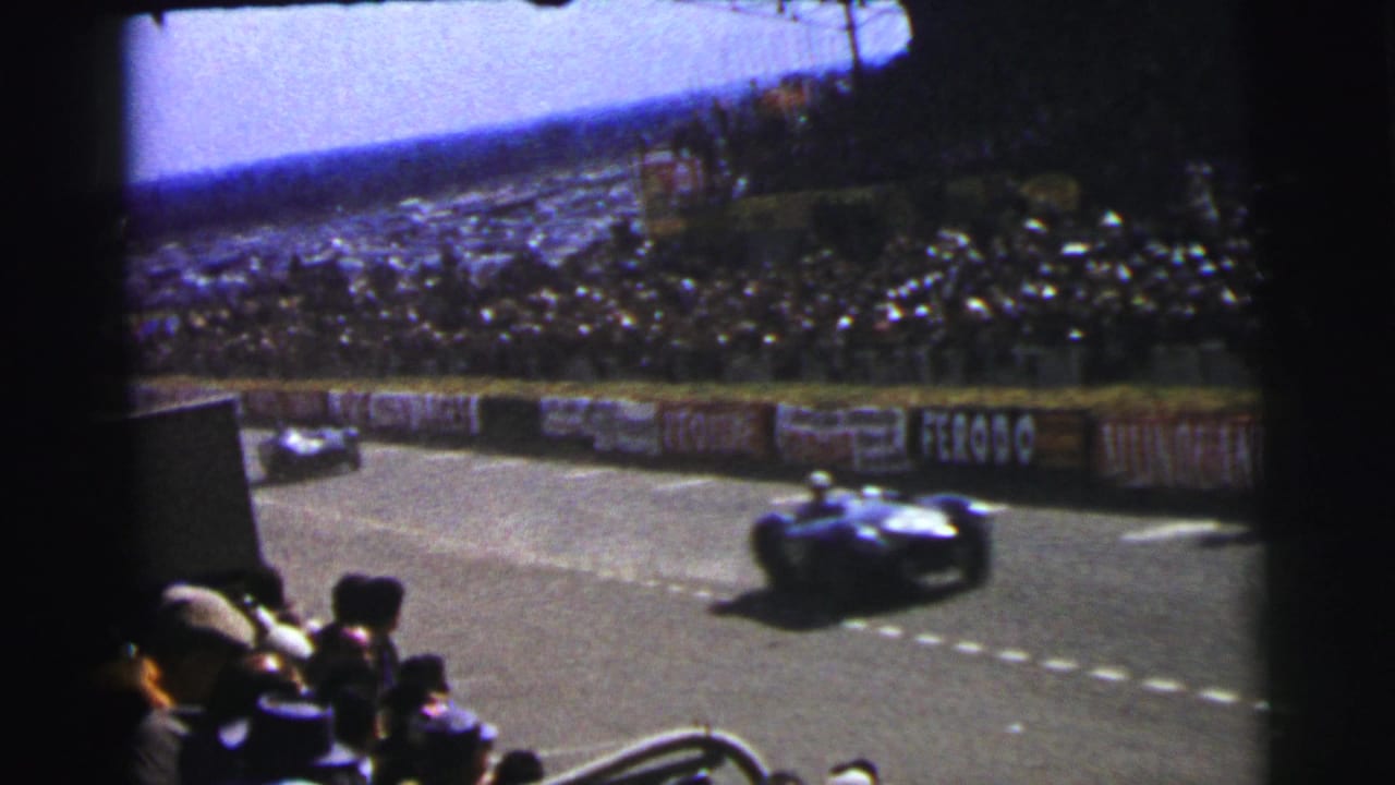 1954 Le Mans frame with one car blurred near the foreground, another farther back, spectators, stands, and visible advertising boards.