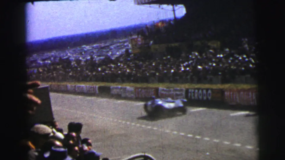 1954 Le Mans spectator view of a race car speeding past, with trackside boards, foreground viewers, and a dense crowd behind the barrier.