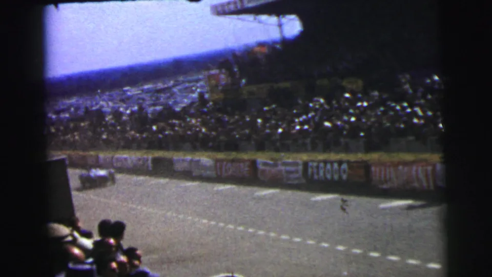 1954 Le Mans frame showing a race car receding along the circuit, foreground spectators, trackside advertising, and packed stands under bright sky.