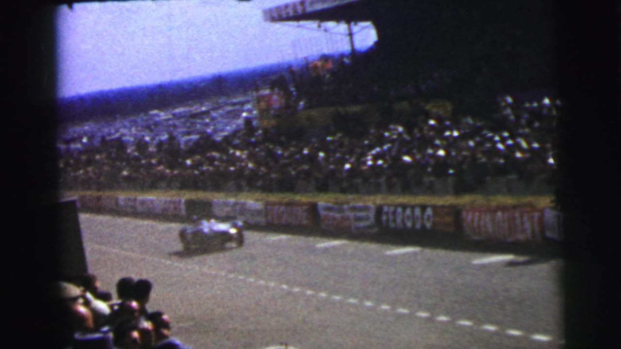 Open-wheel race car passes a crowded circuit grandstand with sponsor boards along the barrier.