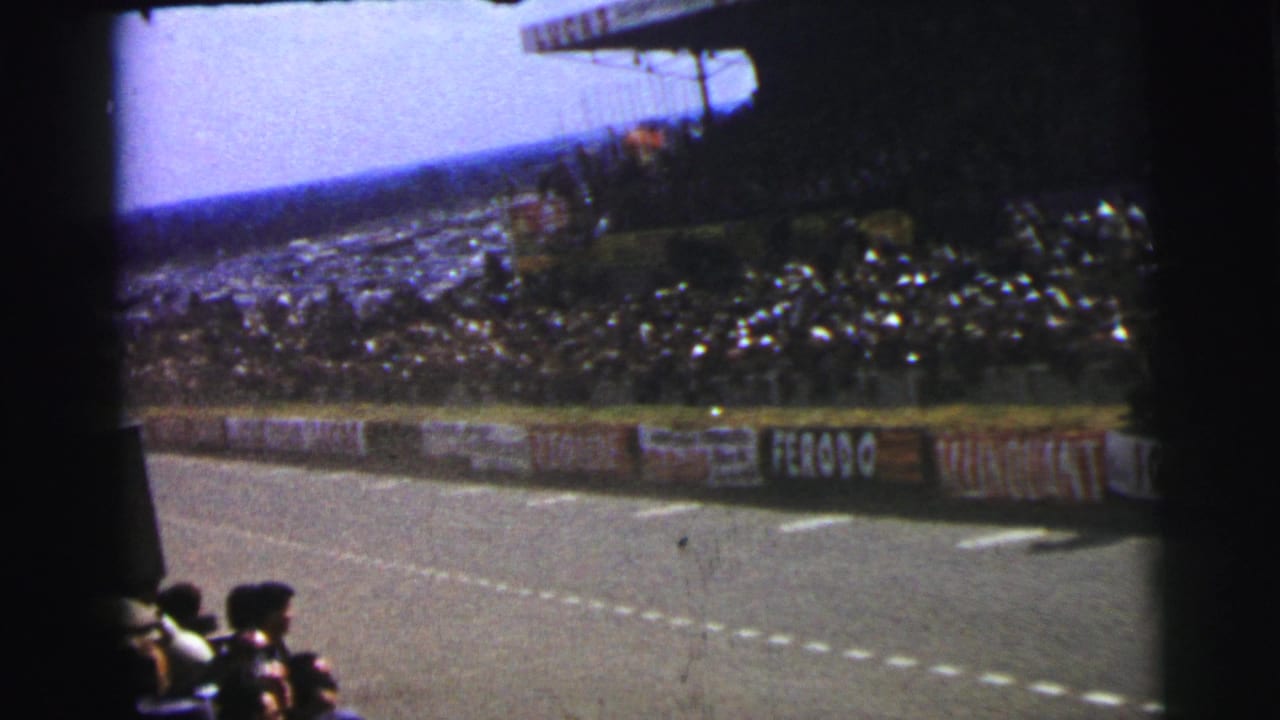 1954 Le Mans trackside view showing the circuit, dense spectator stands, nearby seated viewers, and visible sponsor signage.
