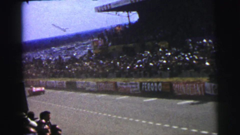 1954 Le Mans spectator footage with a low aircraft overhead, crowded grandstands, trackside banners, and a race car at frame edge.