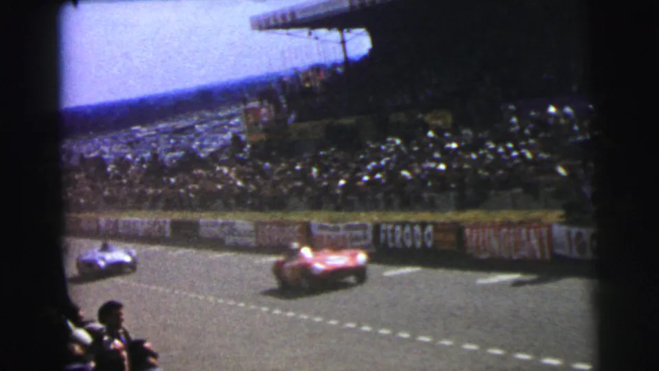 1954 Le Mans frame showing two race cars on the circuit, crowded stands, trackside advertising, and nearby spectators in silhouette.