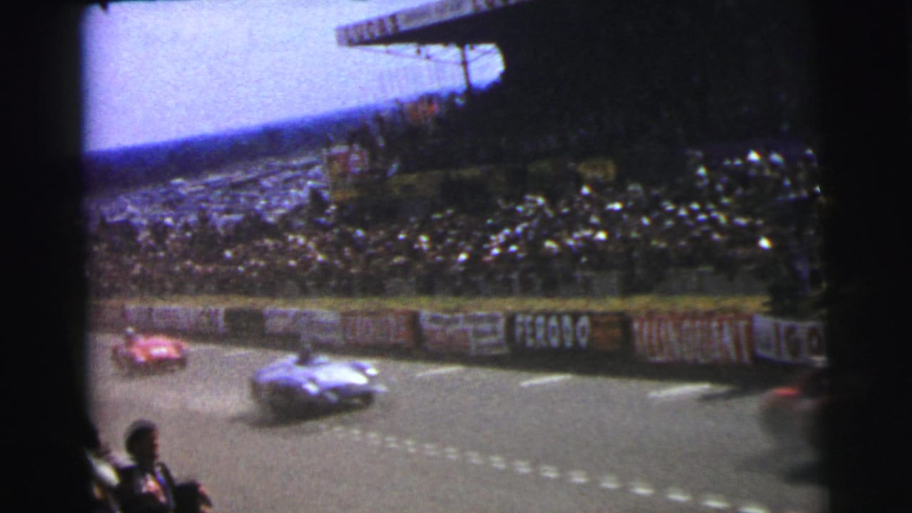 Two race cars pass a crowded grandstand on a paved circuit.