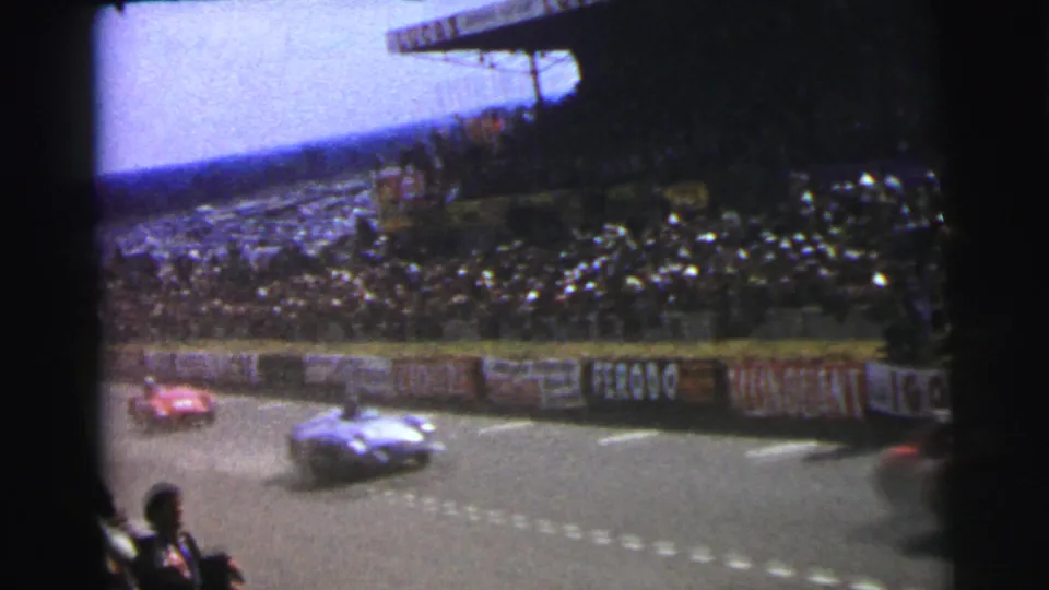 1954 Le Mans home-movie frame showing red and silver race cars passing a packed grandstand beside advertising-lined barriers.