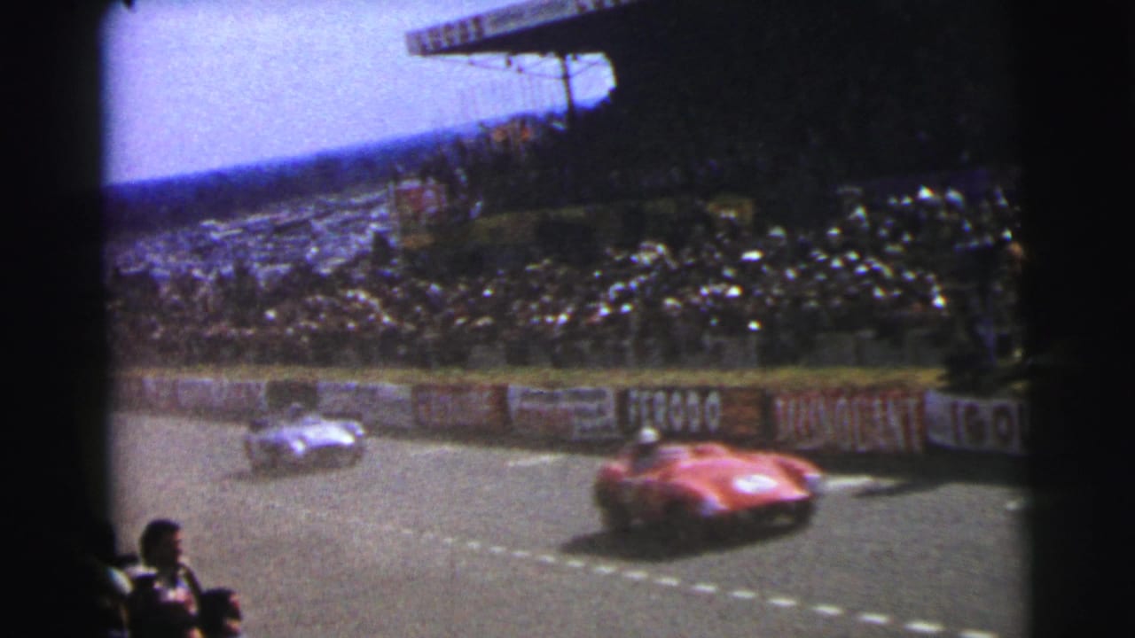 1954 Le Mans frame with a red sports car nearest the camera, a silver car ahead, and spectators packed into the grandstand.
