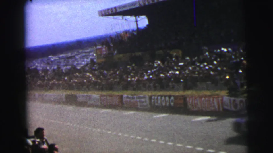 1954 Le Mans home-movie frame after the cars pass, showing the circuit edge, packed stand, and visible barrier advertisements.