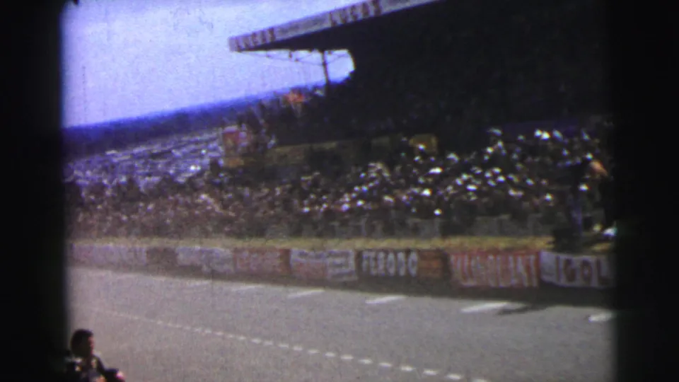 1954 Le Mans frame showing spectators in a shaded grandstand above advertising-lined barriers and the edge of the circuit.