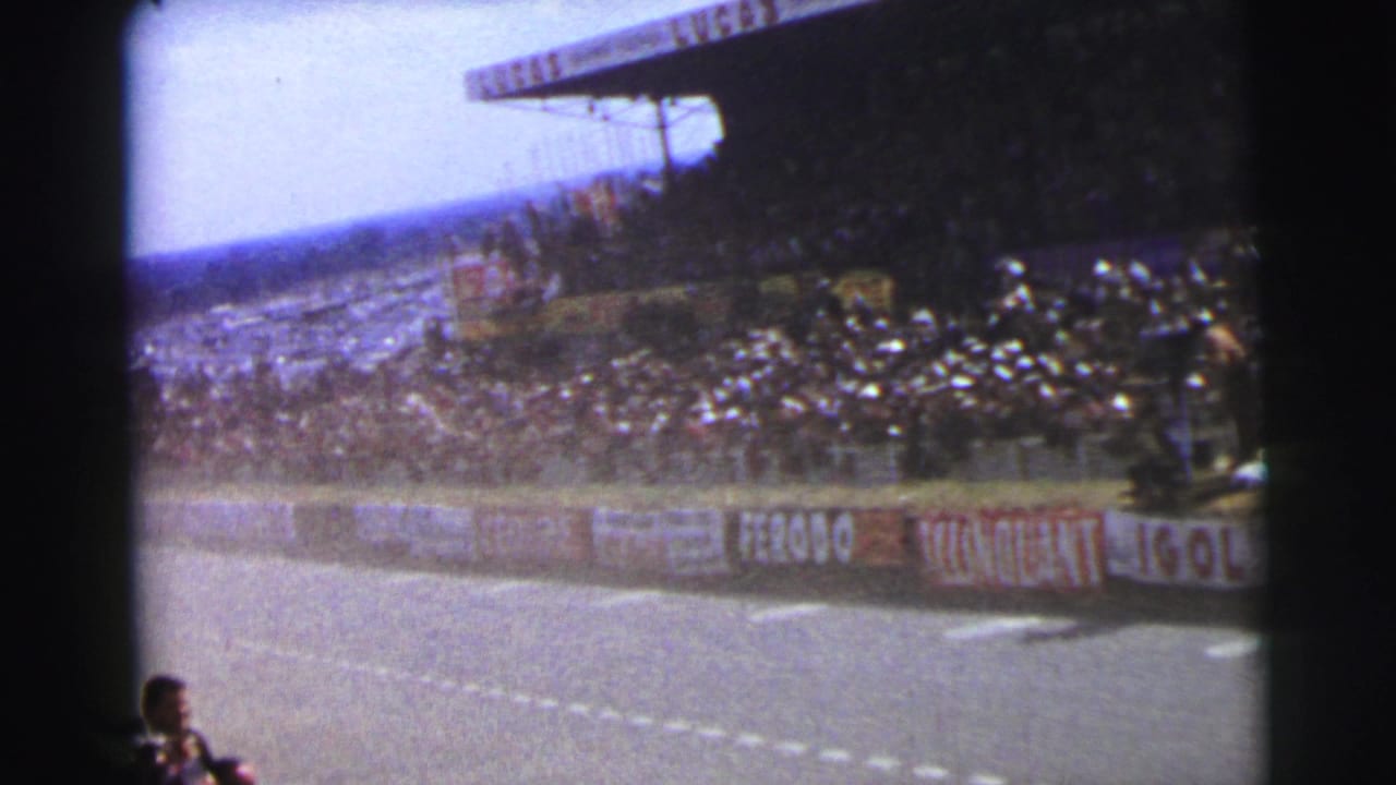 1954 Le Mans 8mm frame showing a race track, crowded grandstand, trackside barriers, and spectators near the camera.