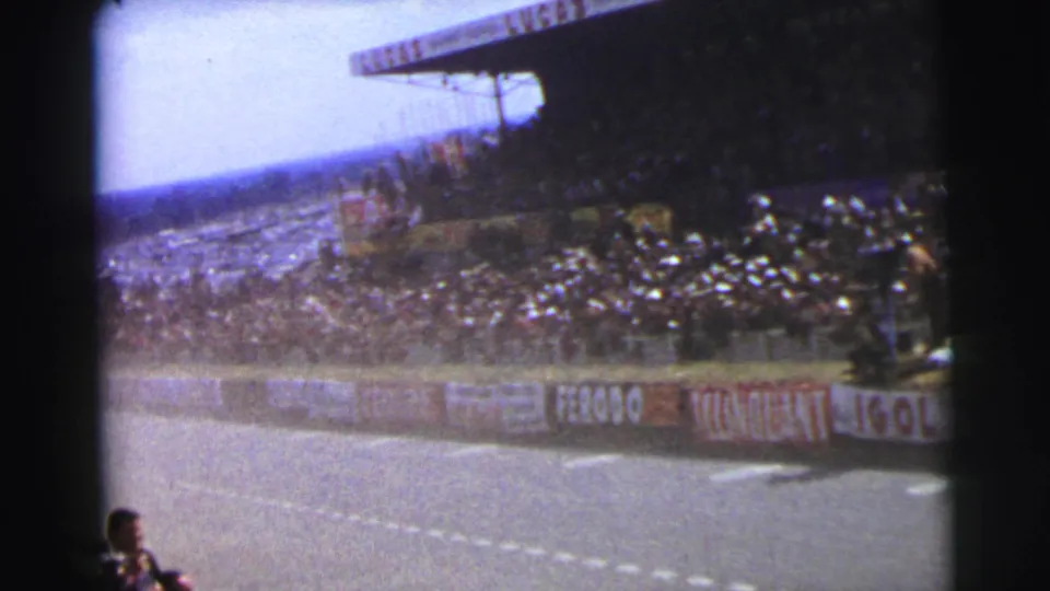 1954 Le Mans 8mm frame showing a race track, crowded grandstand, trackside barriers, and spectators near the camera.