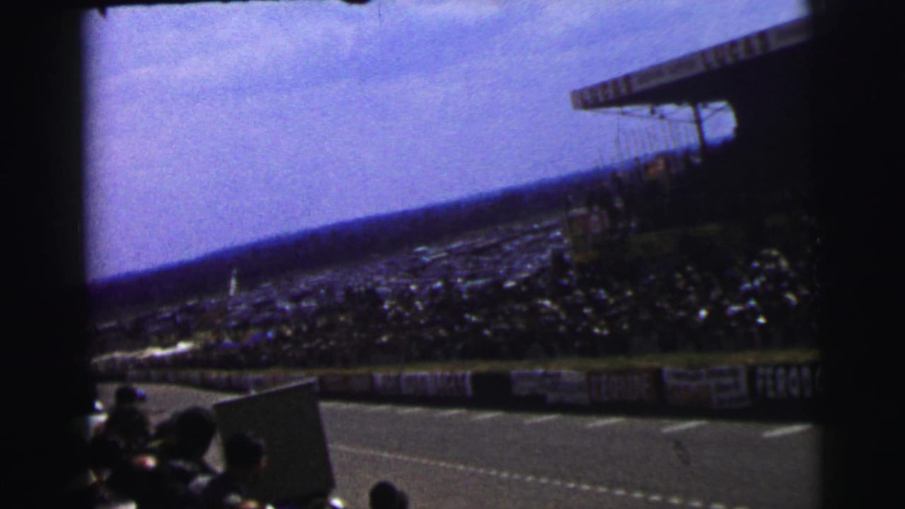Crowded race grandstand beside a paved track with spectators in silhouette at the lower edge.