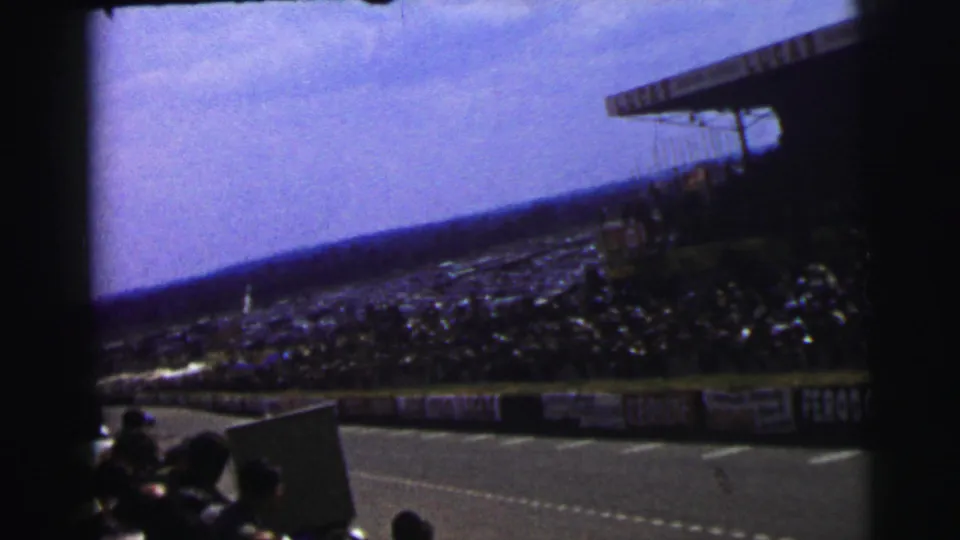 Le Mans trackside view with a large spectator stand, advertising boards, open sky, and silhouetted viewers in the foreground.
