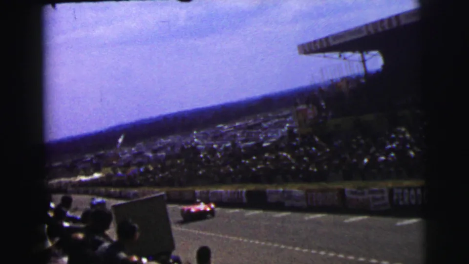 1954 Le Mans spectator footage catches a red racing car on the track below the grandstand and advertising barriers.