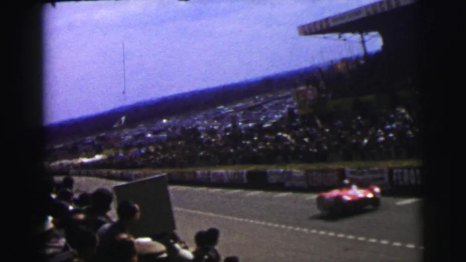 Le Mans 1954 8mm frame showing a red race car passing trackside spectators, advertising boards, and a full grandstand.