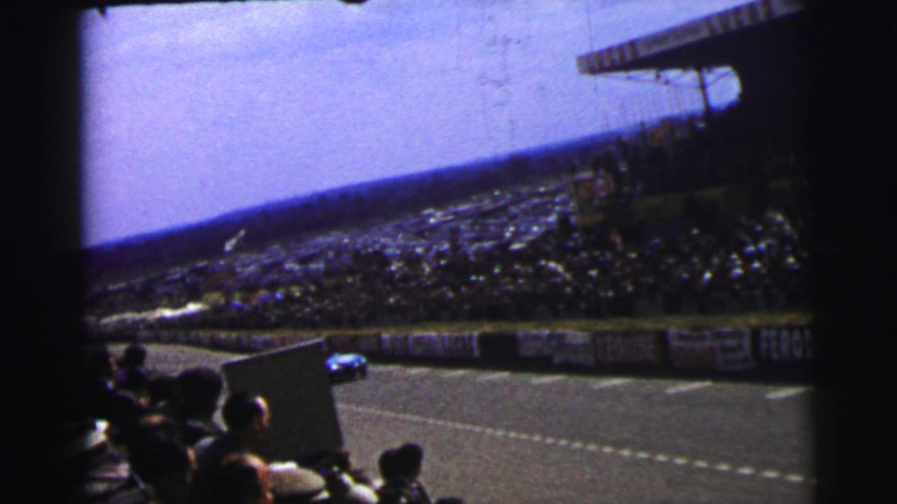 1954 Le Mans home-movie view from the stands, showing spectators, track barriers, and a blue racing car in the distance.