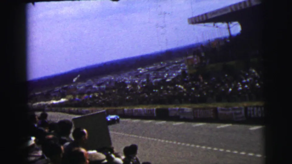 1954 Le Mans home-movie view from the stands, showing spectators, track barriers, and a blue racing car in the distance.